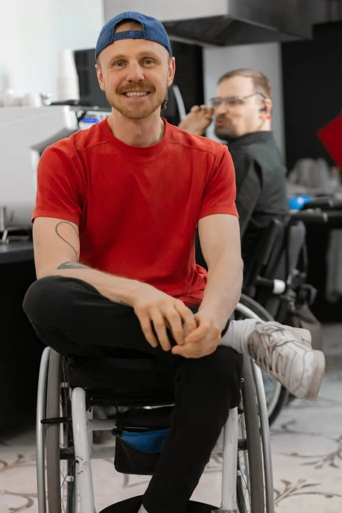 pexels-photo-7698351-7698351 A happy man wearing a red t-shirt and baseball cap smiles while sitting in a wheelchair indoors.